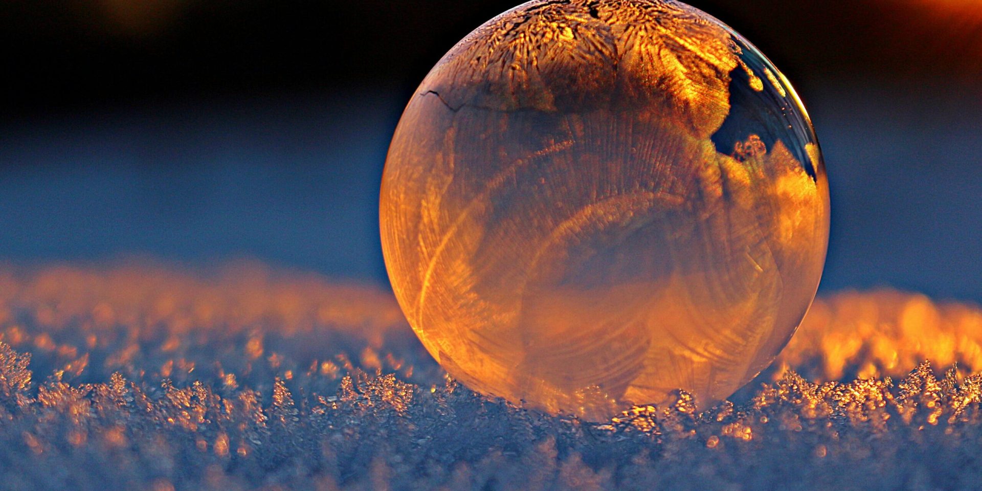 Close-up shot of a frozen bubble with warm reflections resting on a snowy surface at twilight.