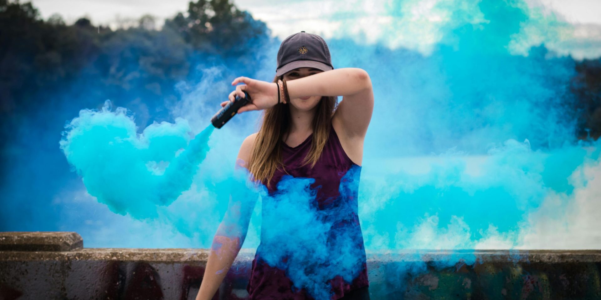 A stylish woman surrounded by bright blue smoke outdoors, creating a dynamic fashion statement.